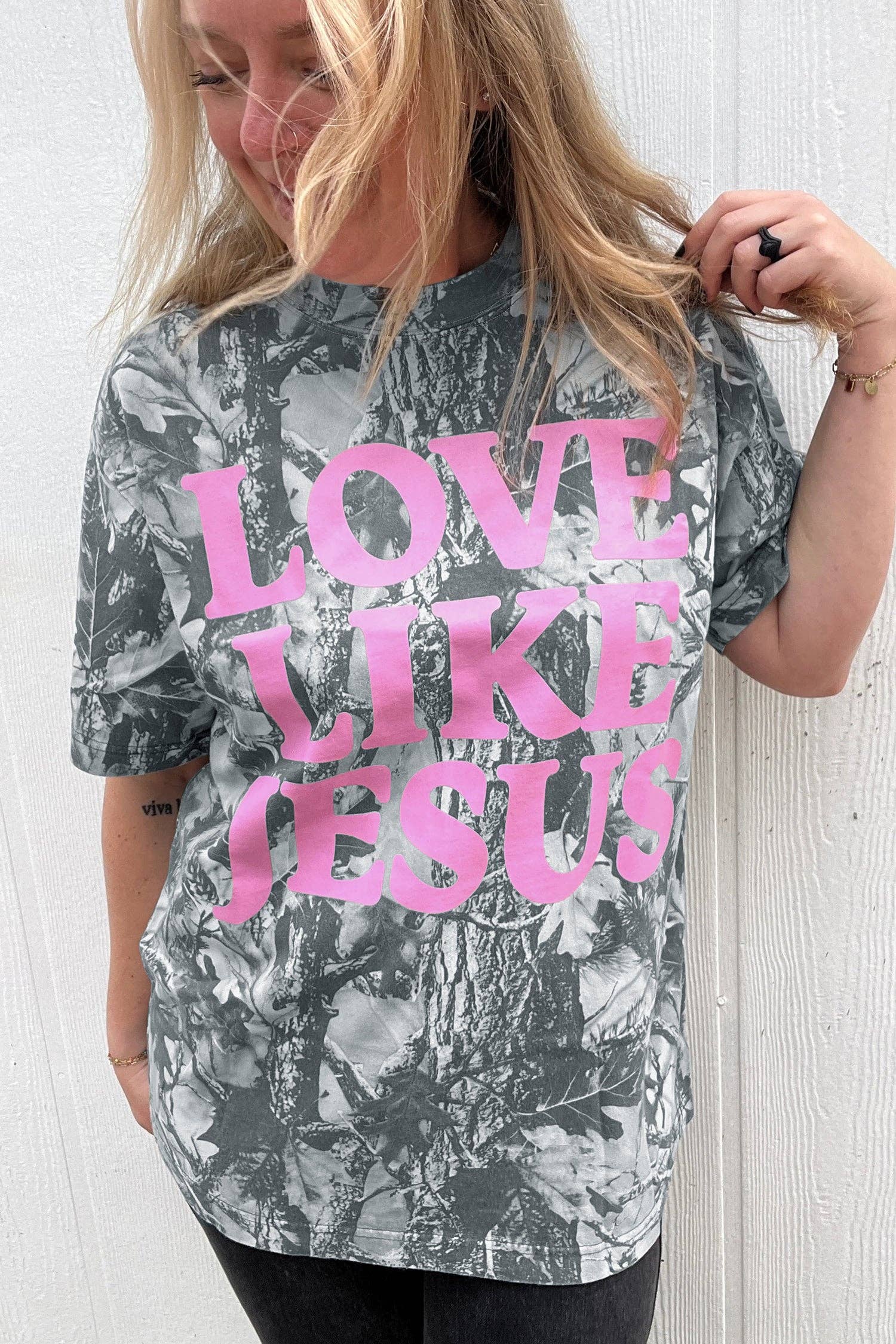 Person wearing a t-shirt with 'LOVE LIKE JESUS' text in pink on a camouflage pattern.