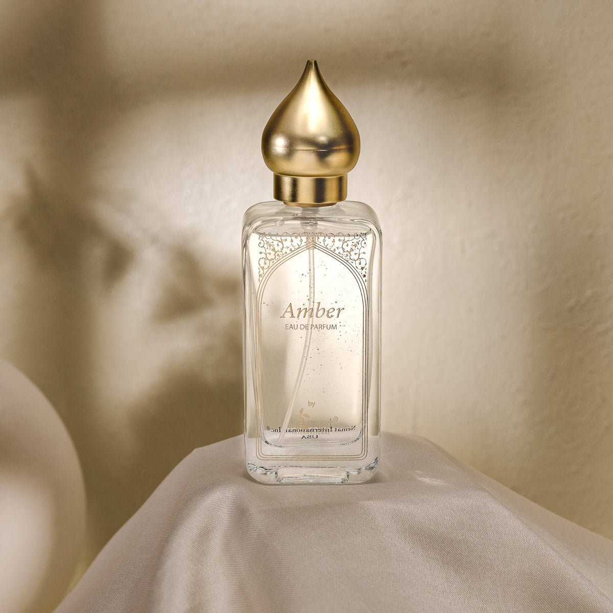 A rectangular glass perfume bottle labeled 'Amber Eau de Parfum' with a metallic gold teardrop-shaped cap, set on draped white fabric against a softly lit beige wall.