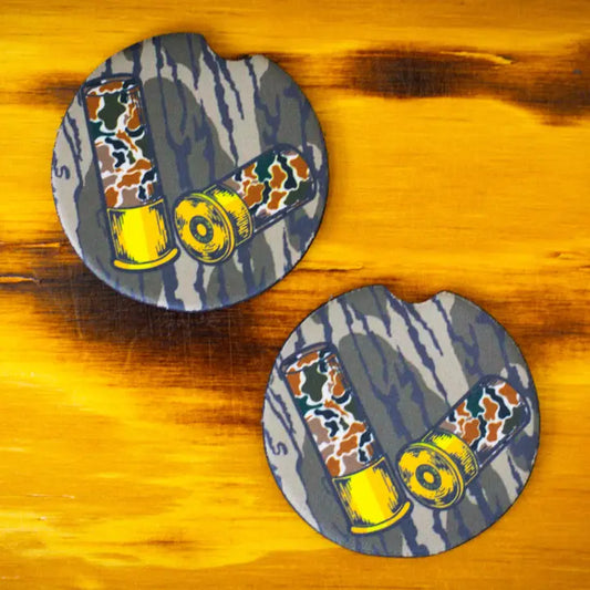 Two round coasters with a gray and blue camouflage pattern, featuring illustrations of shotgun shells in yellow, orange, and brown camouflage, displayed on a yellow wooden surface.