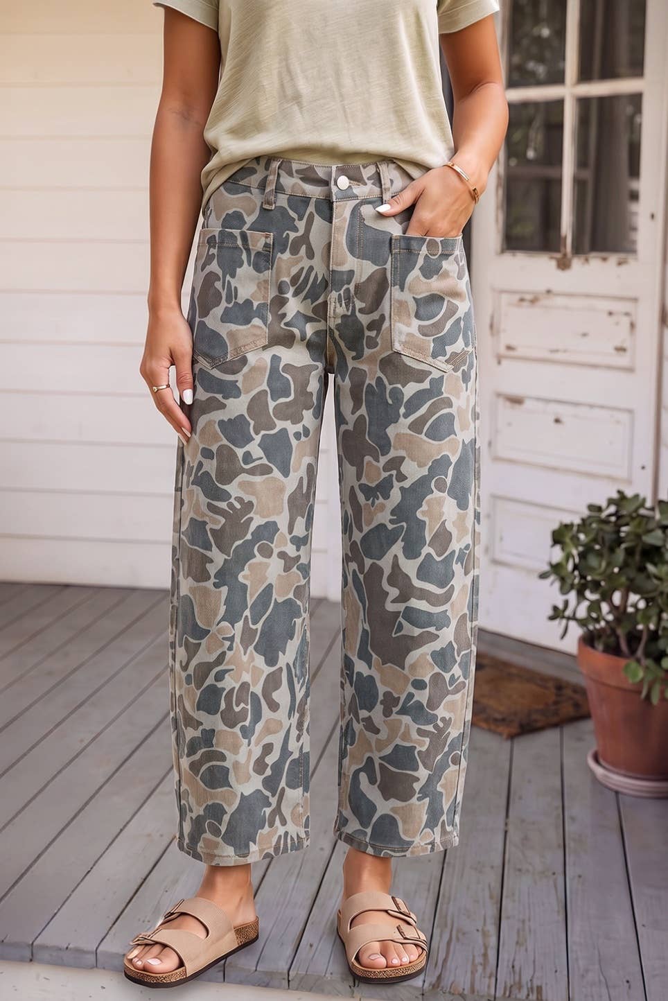 A person wearing loose, wide-leg cropped pants with a muted camouflage pattern in beige, gray, and brown, paired with a light olive top and tan slide sandals, standing on a wooden deck in front of a white porch door.