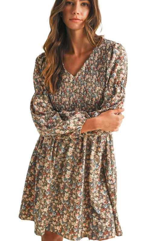 A woman wearing a long-sleeve V-neck dress with a dense small floral print in muted beige, brown, and blue tones, photographed against a plain white background.
