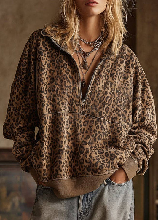 A woman wearing a brown and black leopard print half-zip pullover hoodie with a relaxed fit, styled with layered silver necklaces and light-wash jeans.